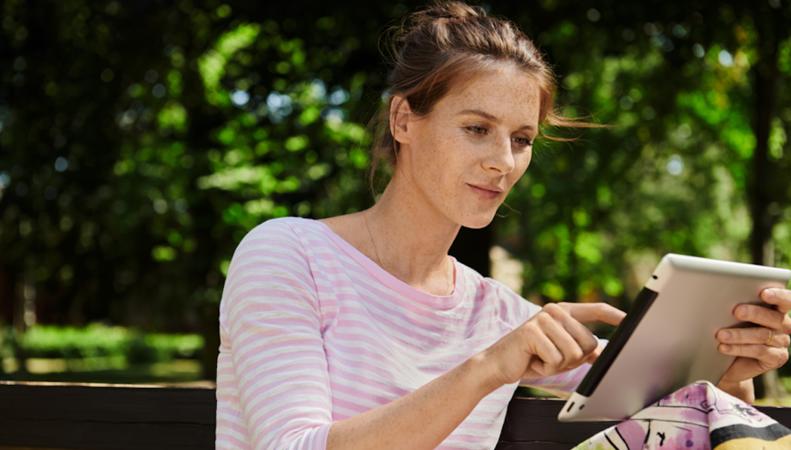 Eine Frau sitzt draußen und hält ein Tablet in der Hand.